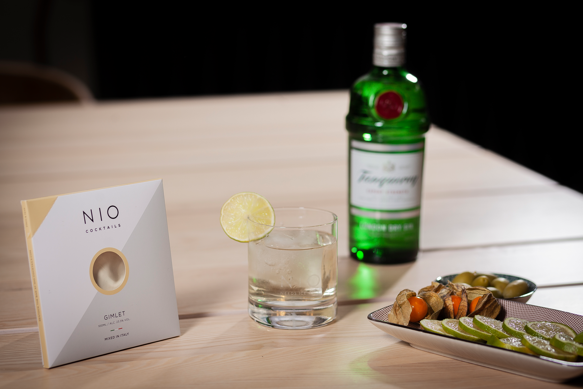 tavolo con box gimlet nio cocktail al gin con bicchiere e bottiglia tanquerey sfocata su sfondo